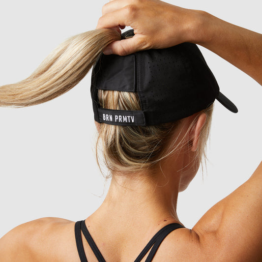 Person wearing a black cap with 'BRN PRMTV' branding, holding their hair back.