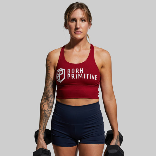 A woman wearing a red sports bra with the brand name 'Born Primitive' on the front, holding dumbbells and standing against a white background.