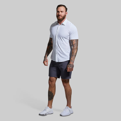 A man wearing a white short sleeve button-down shirt and dark shorts against a plain background